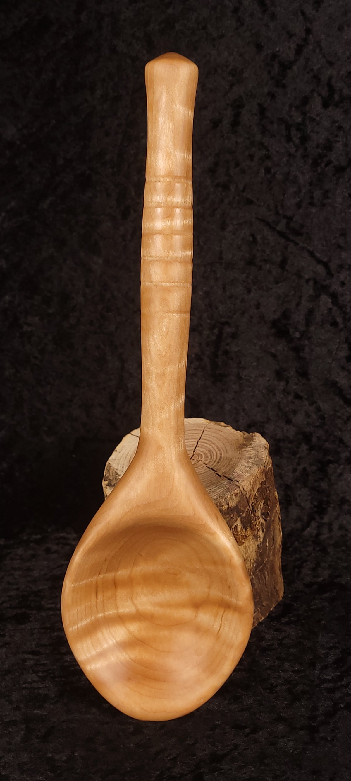 Hand-turned curly maple spoon resting on wood round, back view showing handle rings
